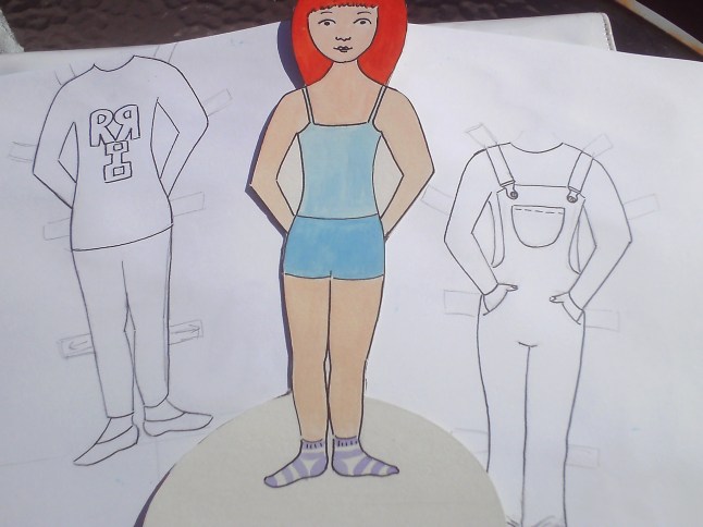 Now draw her some clothes.  I chose to draw her Reflecto Girl costume and her dungarees which she wore over her costume in episode 3 - but you can make any clothes you like, and as many as you like.  Place the cardboard doll on a piece of paper and draw round her.  You can hold them up on a window if you're having difficulty seeing through your paper (this will also help with seeing where the neck and feet are).  Once you have the outlines drawn, you can use your imagination in drawing the clothes.